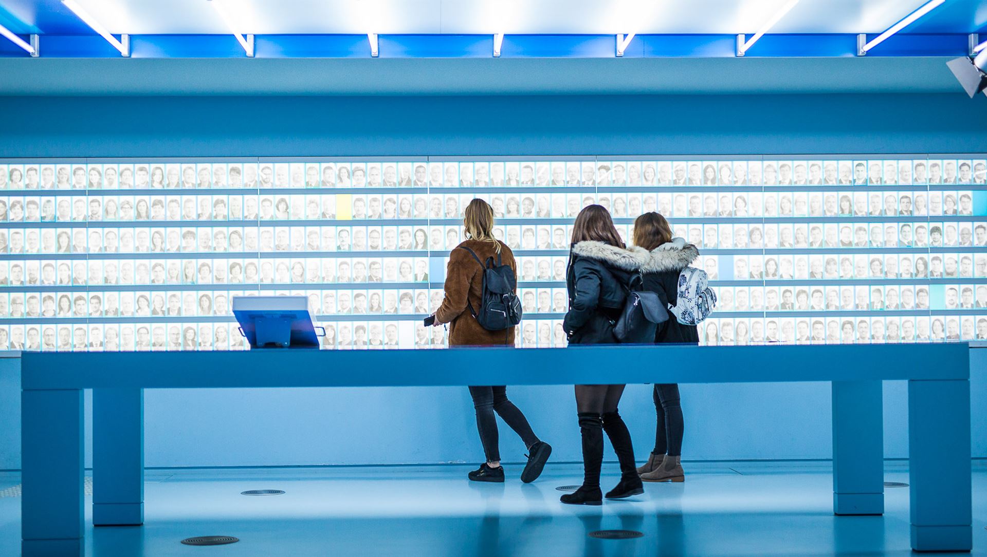 Three young girls looking at hundreds of images of faces on a wall